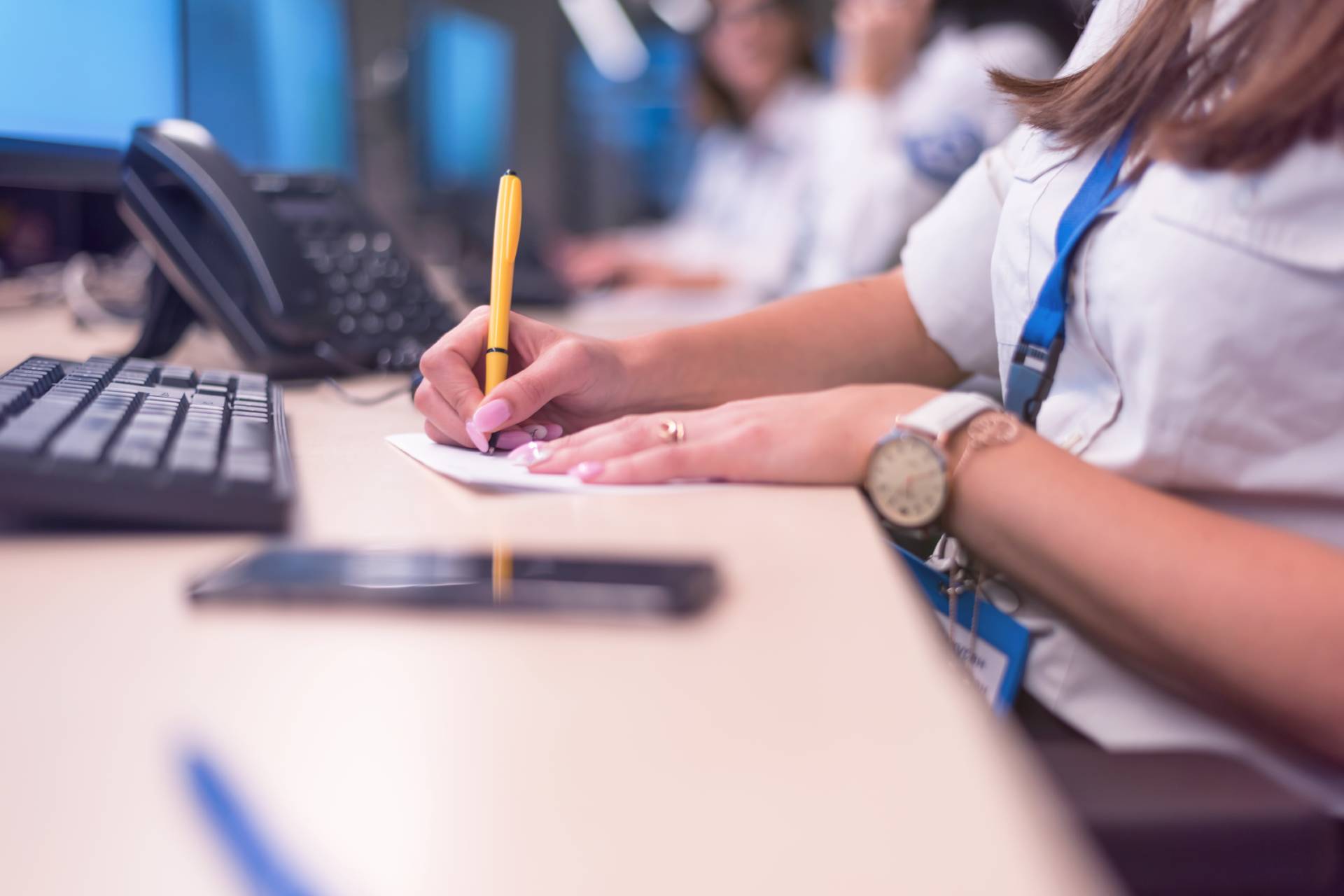 A woman in uniform writing on a notepad at a desk with a keyboard, phone, and computer