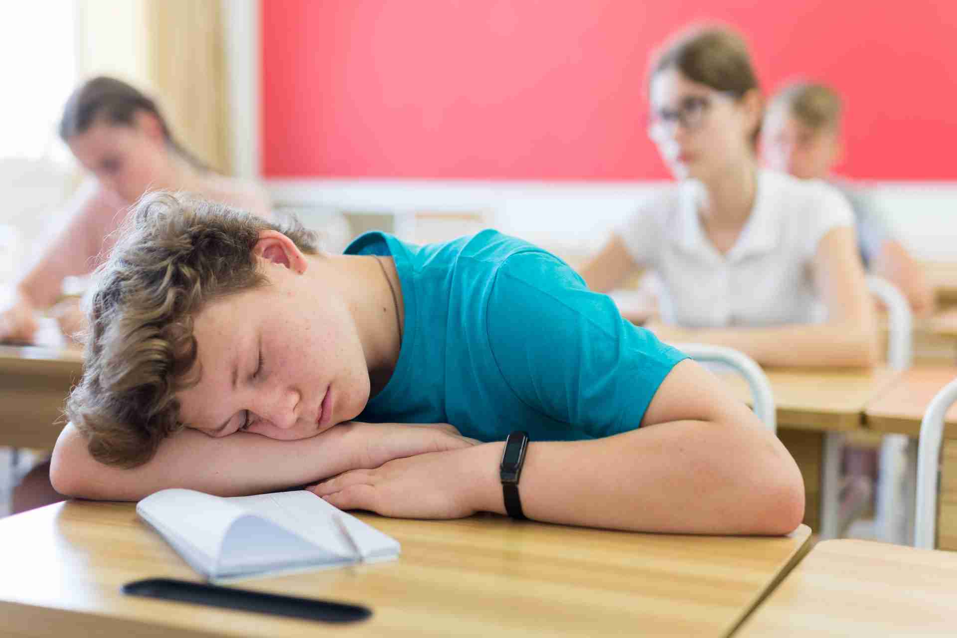 A school student resting their head on a desk with a notebook open, appearing tired during class.
