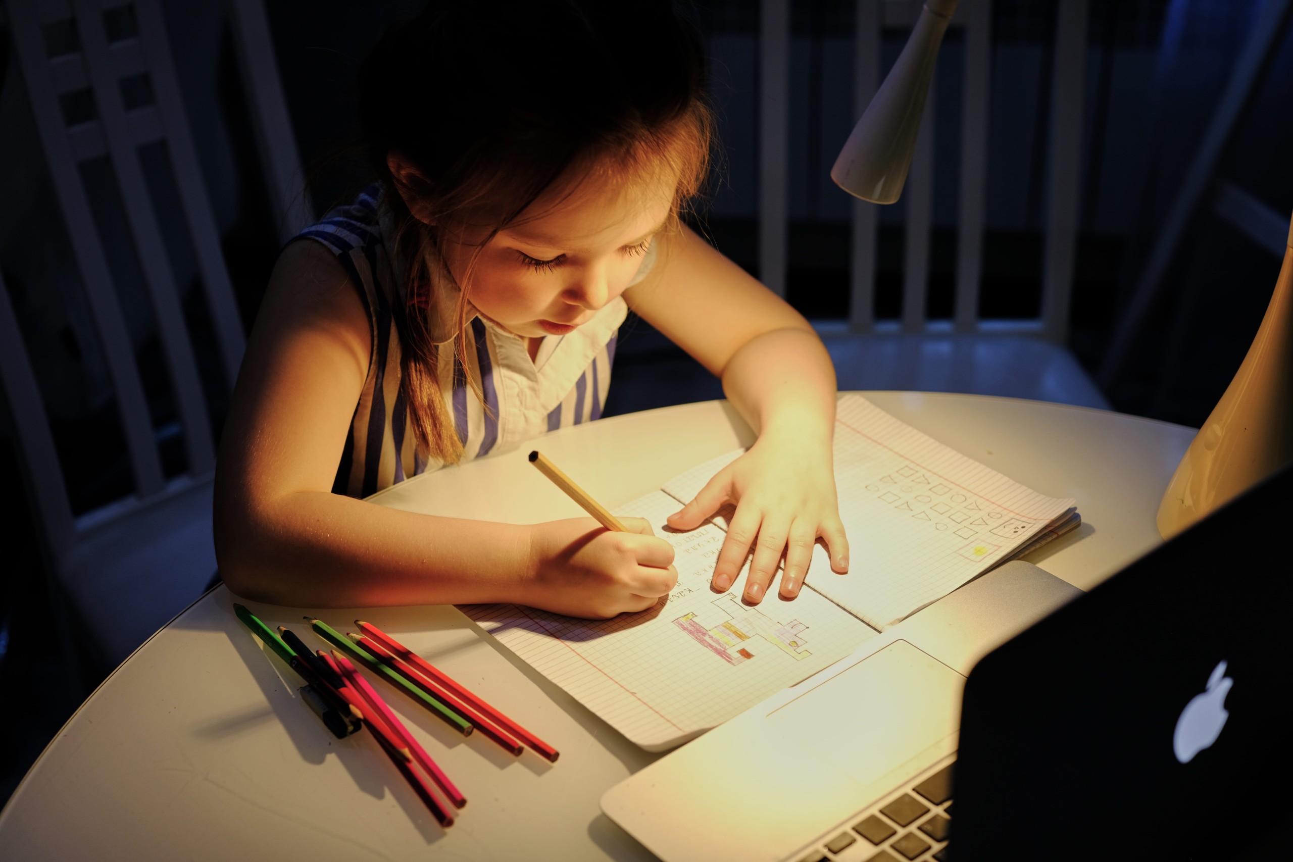 Young child writing in a notebook beside a laptop under warm desk lighting