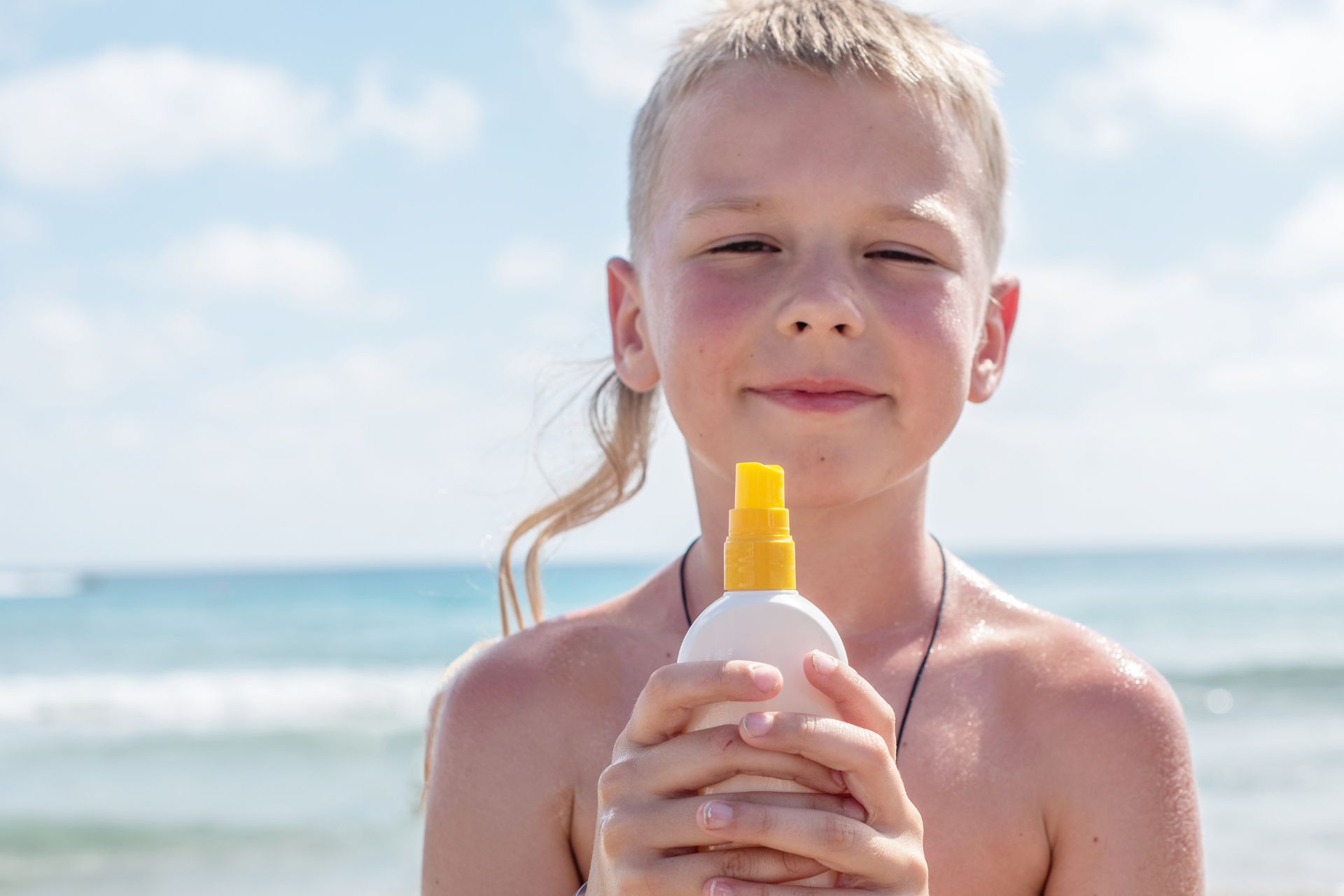 Child holding a bottle of sunscreen outdoors on a sunny day.